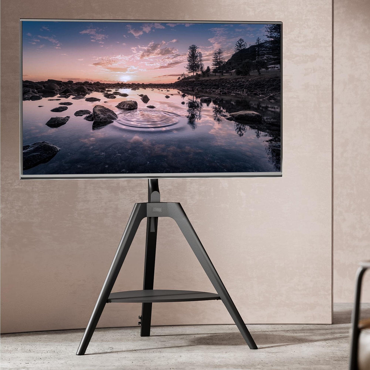 A black tripod TV stand with a shelf, featuring a mounted TV displaying a nature scene. The stand is positioned on a floor with a wall in the background.